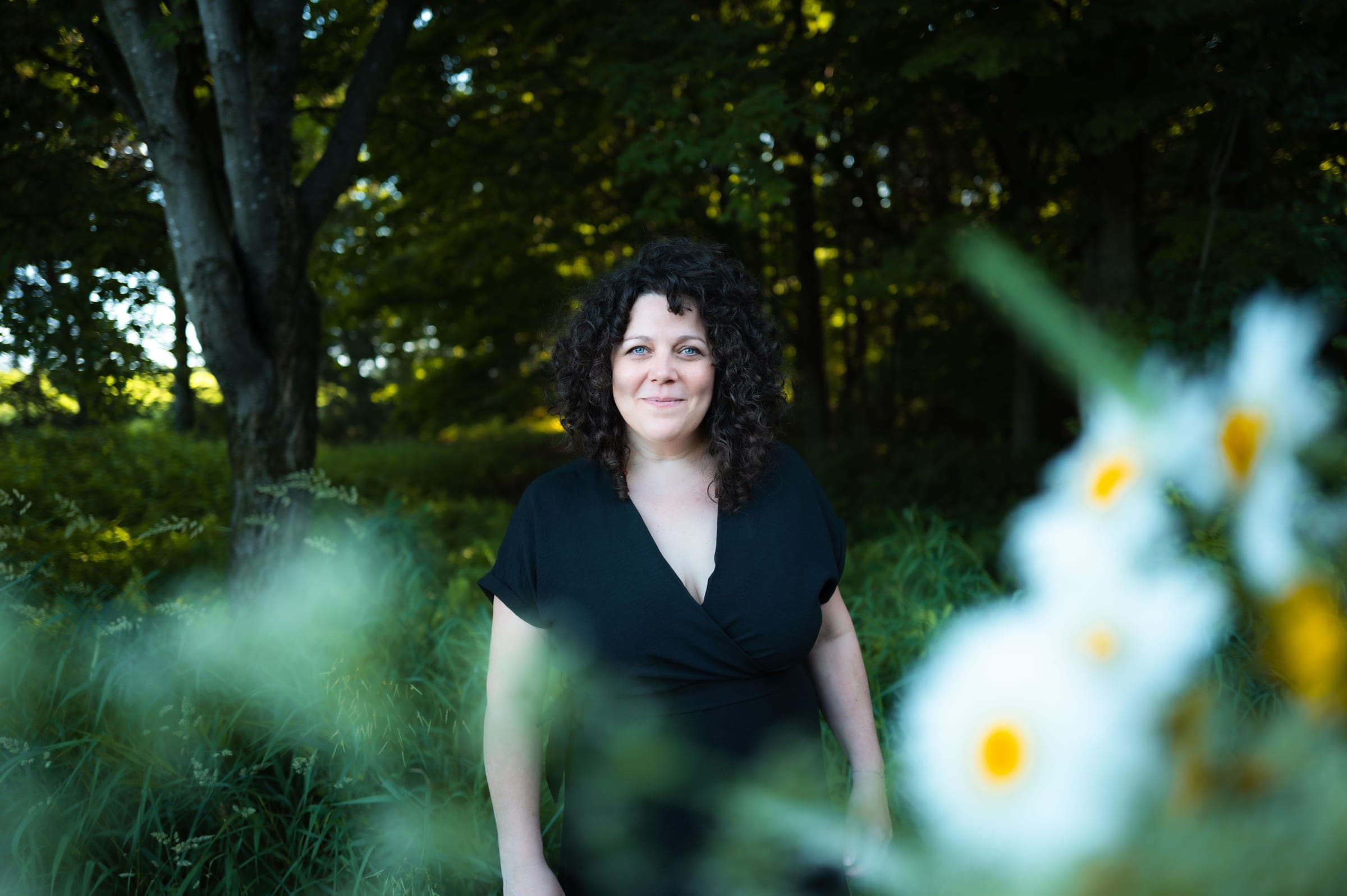 Karine Champagne sourit en robe noire, dans une clairière ombragée, des marguerites floues au premier plan.