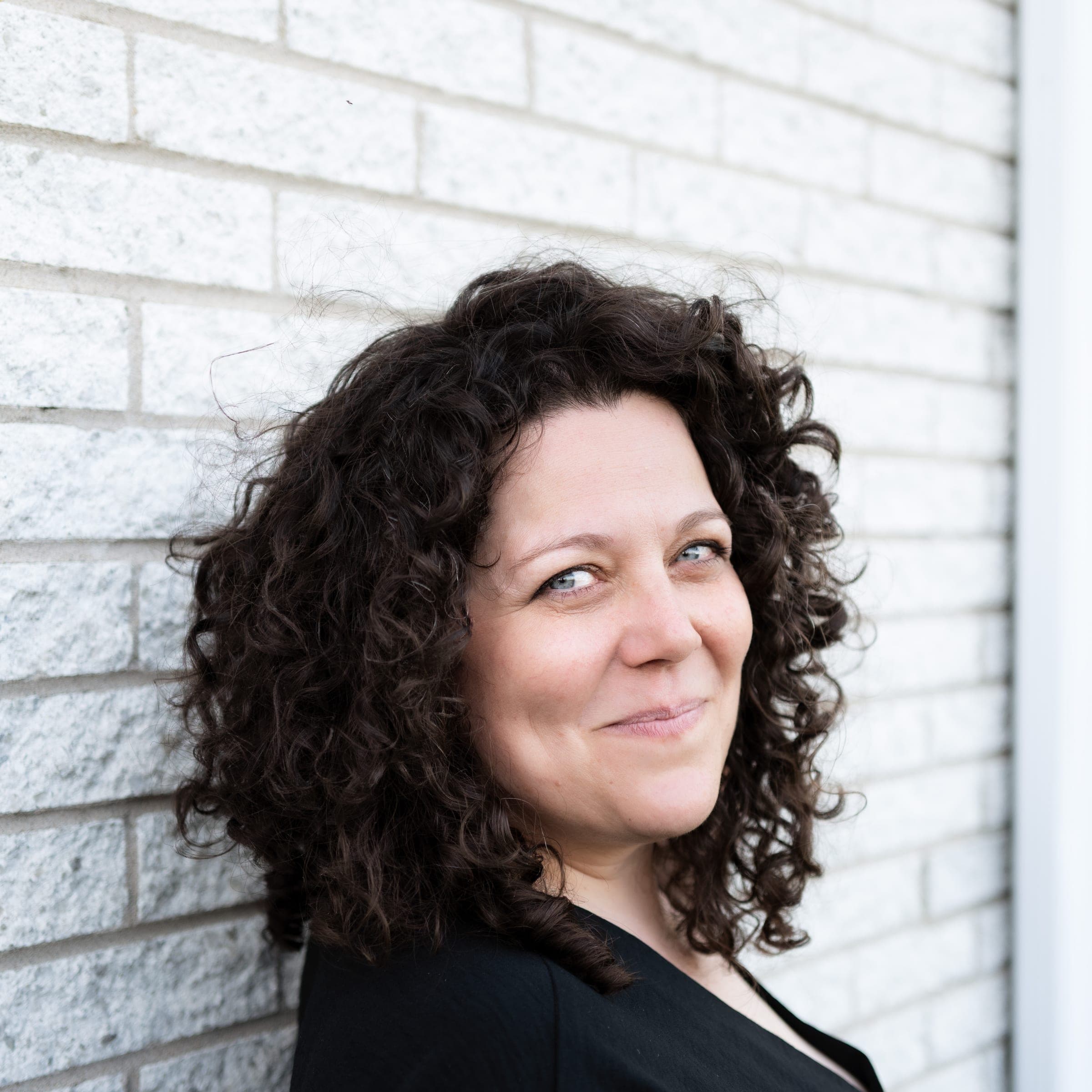 Portrait de Karine Champagne devant un mur de briques blanches, souriante, le regard par-dessus l'épaule.